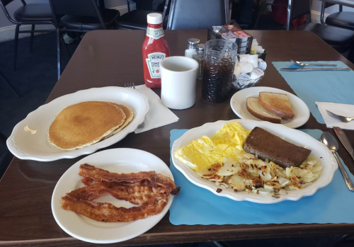 A breakfast spread featuring pancakes, bacon, eggs, hash browns, toast, coffee, and ketchup on a table.
