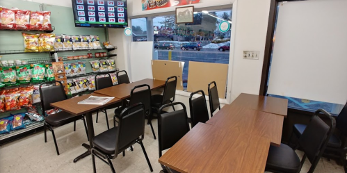 A small dining area with several tables and chairs, snacks on shelves, and a window showing a street view outside.