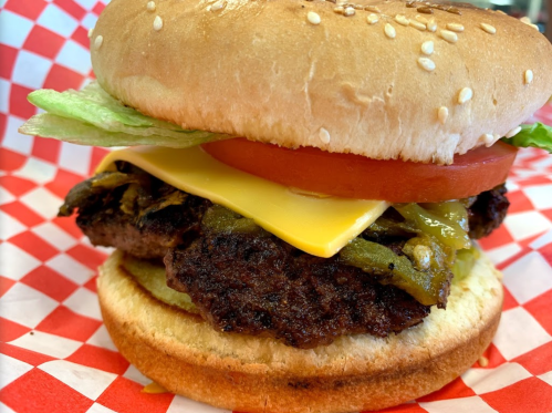 A close-up of a sesame seed bun burger with lettuce, tomato, cheese, and green peppers on a red and white checkered paper.