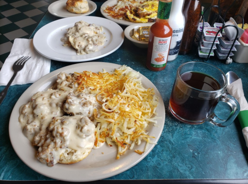 A plate of biscuits and gravy with hash browns, a cup of coffee, and a side of hot sauce on a diner table.