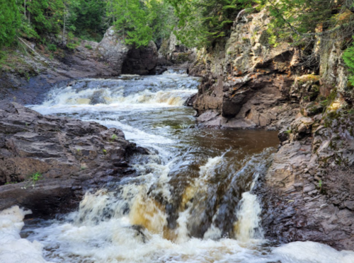 A serene waterfall cascades over rocky terrain, surrounded by lush green trees and a tranquil river.