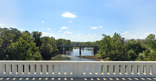 A scenic view of a river with a bridge, surrounded by lush green trees under a clear blue sky.