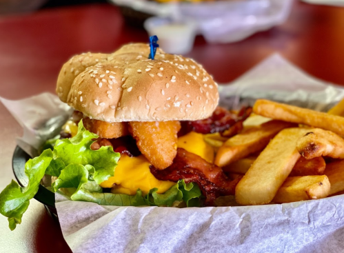 A sesame seed bun burger with lettuce, cheese, bacon, and onion rings, served with a side of French fries.
