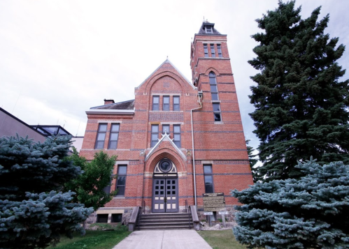 Historic brick building with a tall clock tower, surrounded by green trees and a cloudy sky.