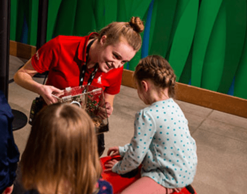 A staff member shows a child a small terrarium while another child watches, in a colorful indoor setting.
