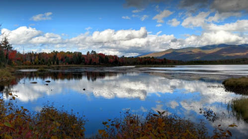 A serene lake reflects colorful autumn trees and a blue sky with clouds, surrounded by mountains in the background.