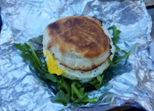A toasted sandwich with egg and greens, wrapped in foil, resting on a wooden surface.