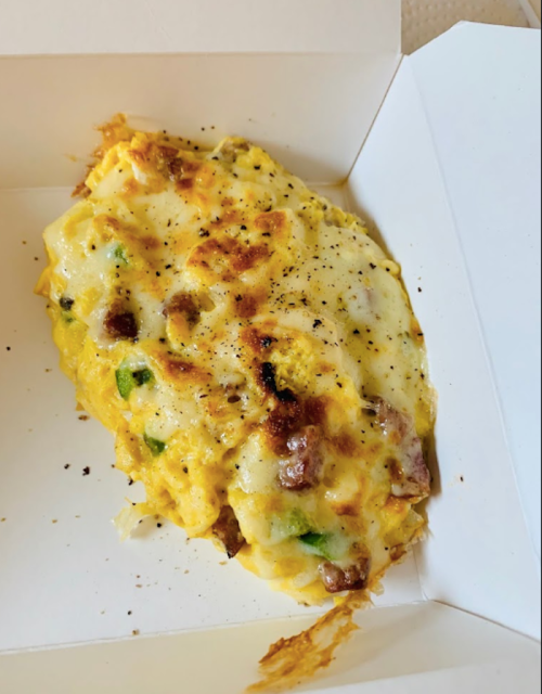 A cheesy, baked egg dish with green peppers and meat, served in a white takeout box.