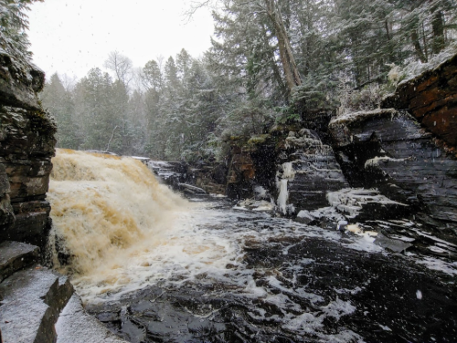 A snowy landscape featuring a waterfall cascading over rocky cliffs into a frothy river below.