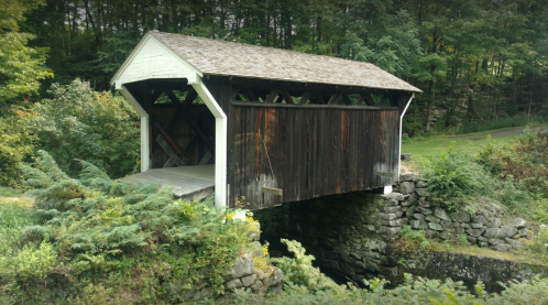 A rustic covered bridge made of wood, resting on stone supports, surrounded by lush greenery and trees.