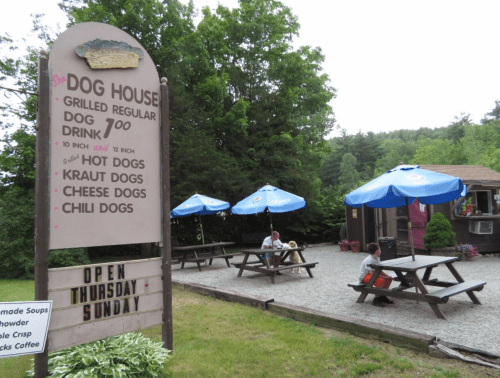 A small eatery called "The Dog House" with outdoor seating, offering various hot dogs and drinks, surrounded by trees.
