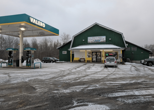 A Valero gas station with a green building, snowy ground, and parked cars in a rural area.