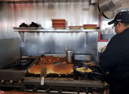 A chef cooking on a griddle with meats and eggs in a busy kitchen, surrounded by plates and cooking utensils.