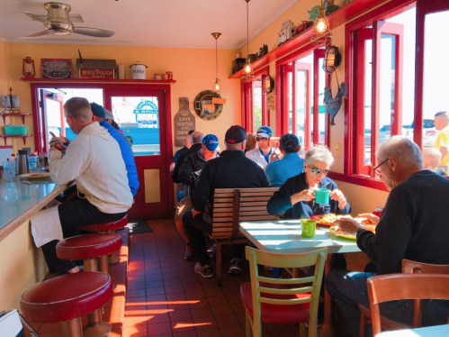 A cozy diner scene with patrons enjoying meals at tables, bright red decor, and large windows letting in sunlight.
