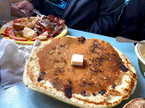 A large pancake with a pat of butter sits in front of a plate of bacon, fruit, and French toast.