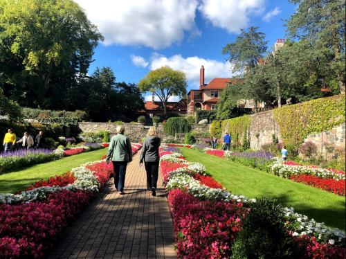 Two people walk down a flower-lined path in a vibrant garden, with lush greenery and a blue sky in the background.