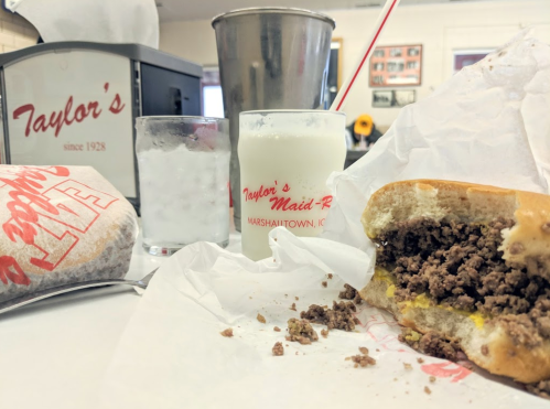 A close-up of a half-eaten sandwich with ground beef, a glass of milk, and a soda in a diner setting.