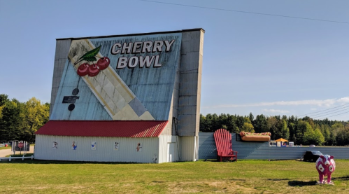 A large, colorful mural of cherries on a building, with a giant red chair and a cow statue in the foreground.