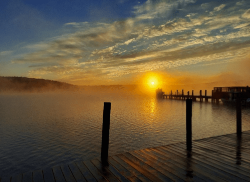 A serene sunrise over a misty lake, with a wooden dock extending into the water and colorful clouds in the sky.