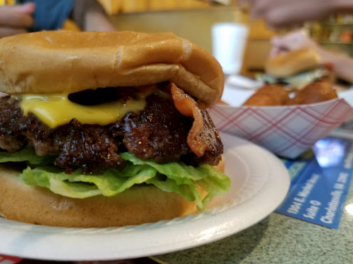 A close-up of a cheeseburger with bacon, lettuce, and a soft bun, with a side of fried snacks in the background.