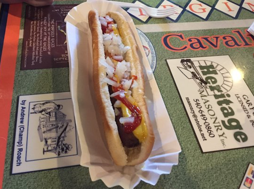 A hot dog in a bun topped with mustard, ketchup, and chopped onions, served on a paper tray.