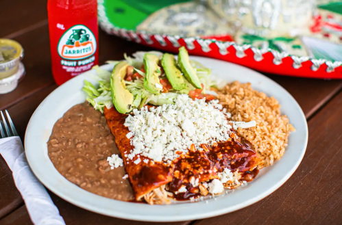 A plate of enchiladas topped with cheese, served with rice, refried beans, and a side of salad, with a drink in the background.