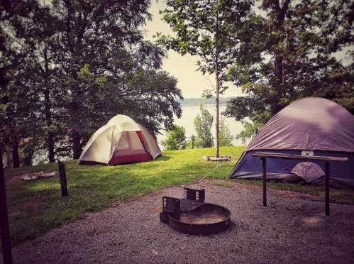 Two tents set up by a lake, surrounded by trees, with a fire pit in the foreground and a peaceful water view.