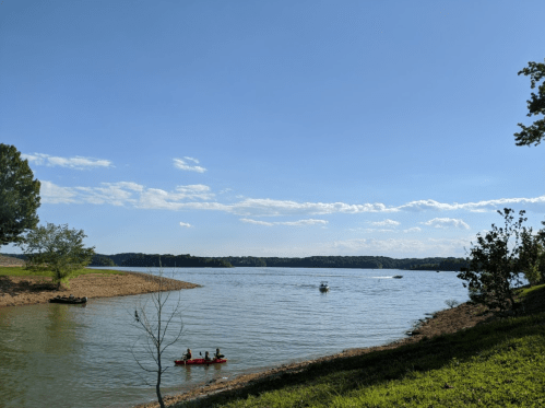 A serene lake scene with two kayakers, blue sky, and lush greenery along the shore.