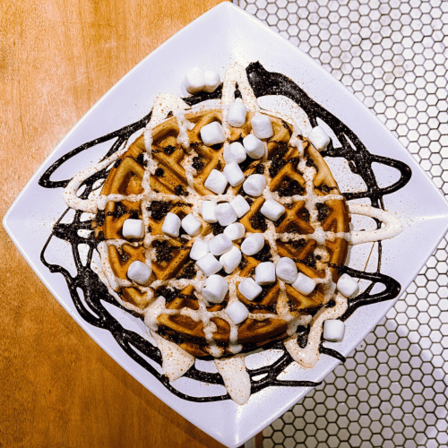 A square plate with a waffle topped with marshmallows and drizzled with chocolate and cream sauces.