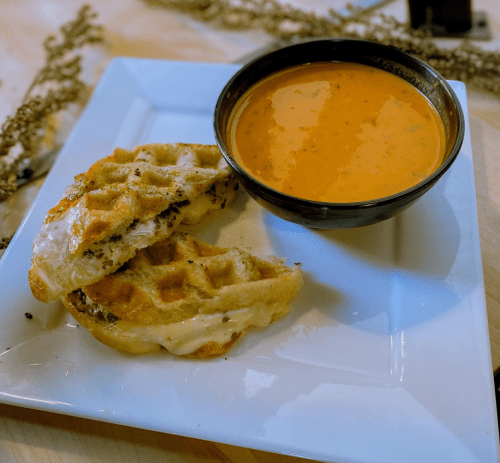 A plate with two grilled cheese waffle sandwiches and a bowl of tomato soup.