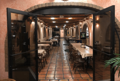 Interior of a restaurant with arched doorways, wooden tables, and chairs, featuring warm lighting and tiled flooring.