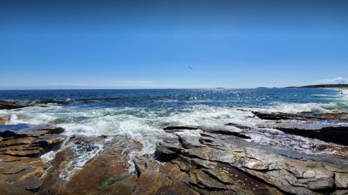 A rocky shoreline with waves crashing against the rocks under a clear blue sky and distant horizon.