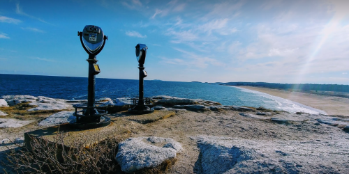 Two coin-operated binoculars on a rocky outcrop overlooking a beach and ocean under a clear blue sky.