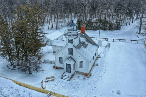 Aerial view of a white lighthouse surrounded by snow-covered trees and a winter landscape.