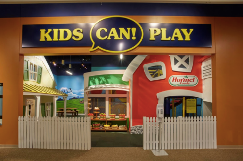 Colorful play area entrance with a sign "KIDS CAN! PLAY" and a white picket fence, featuring a red and green building design.