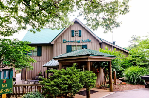 A rustic building with a green roof and wooden exterior, surrounded by trees and shrubs, featuring a sign that reads "Banning Mills."