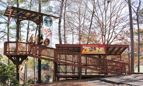 Wooden zipline platform with signs in a forested area, featuring a staircase and a welcoming banner.
