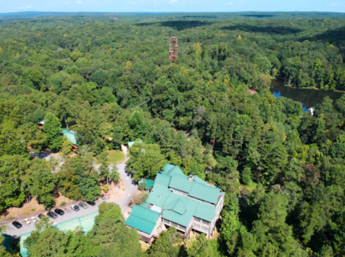 Aerial view of a green forest with a large building and a pond, surrounded by trees and a lookout tower in the distance.