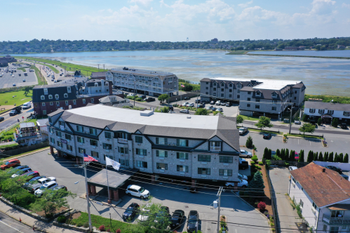 Aerial view of a hotel near a river, with cars parked and a highway visible in the background under a blue sky.