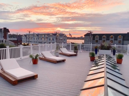 A rooftop deck with lounge chairs overlooks a sunset and buildings by the water, creating a serene atmosphere.