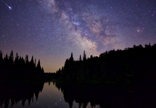 A serene night sky filled with stars and the Milky Way, reflecting on a calm lake surrounded by dark silhouettes of trees.
