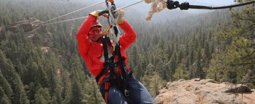 A person in a red jacket zip-lining over a forested area, gripping the harness with trees in the background.