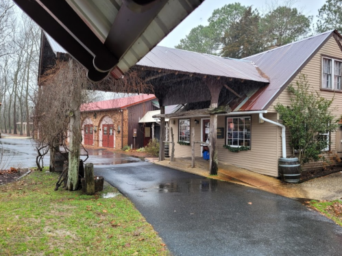 A rustic building with a covered porch, surrounded by trees, on a rainy day. Wet pavement reflects the overcast sky.