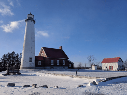 A tall lighthouse beside a red-roofed house and a small shed, surrounded by snow under a clear blue sky.