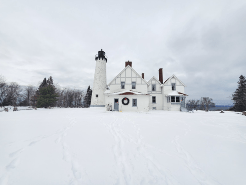 A white lighthouse beside a snowy house, surrounded by trees under a cloudy sky.
