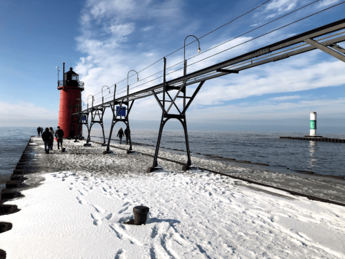 A red lighthouse on a snowy pier, with people walking along the icy surface and a green buoy in the distance.