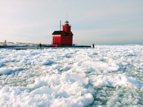 A red lighthouse stands on a pier surrounded by ice and snow, with a calm sky in the background.