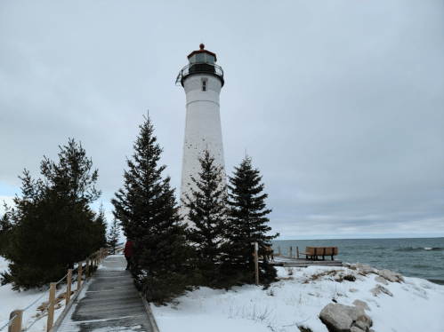 A tall white lighthouse surrounded by snow-covered trees, near a rocky shoreline and calm blue waters under a cloudy sky.