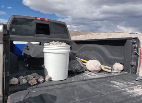 A pickup truck bed filled with rocks, a white bucket, and a yellow tool, set against a cloudy sky.