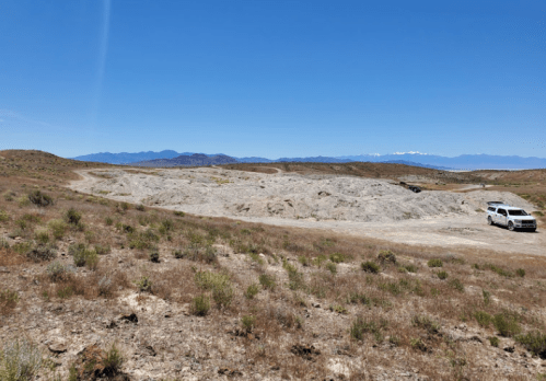 A barren landscape with sparse vegetation, a white car parked on a dirt road, and mountains in the distance under a clear blue sky.